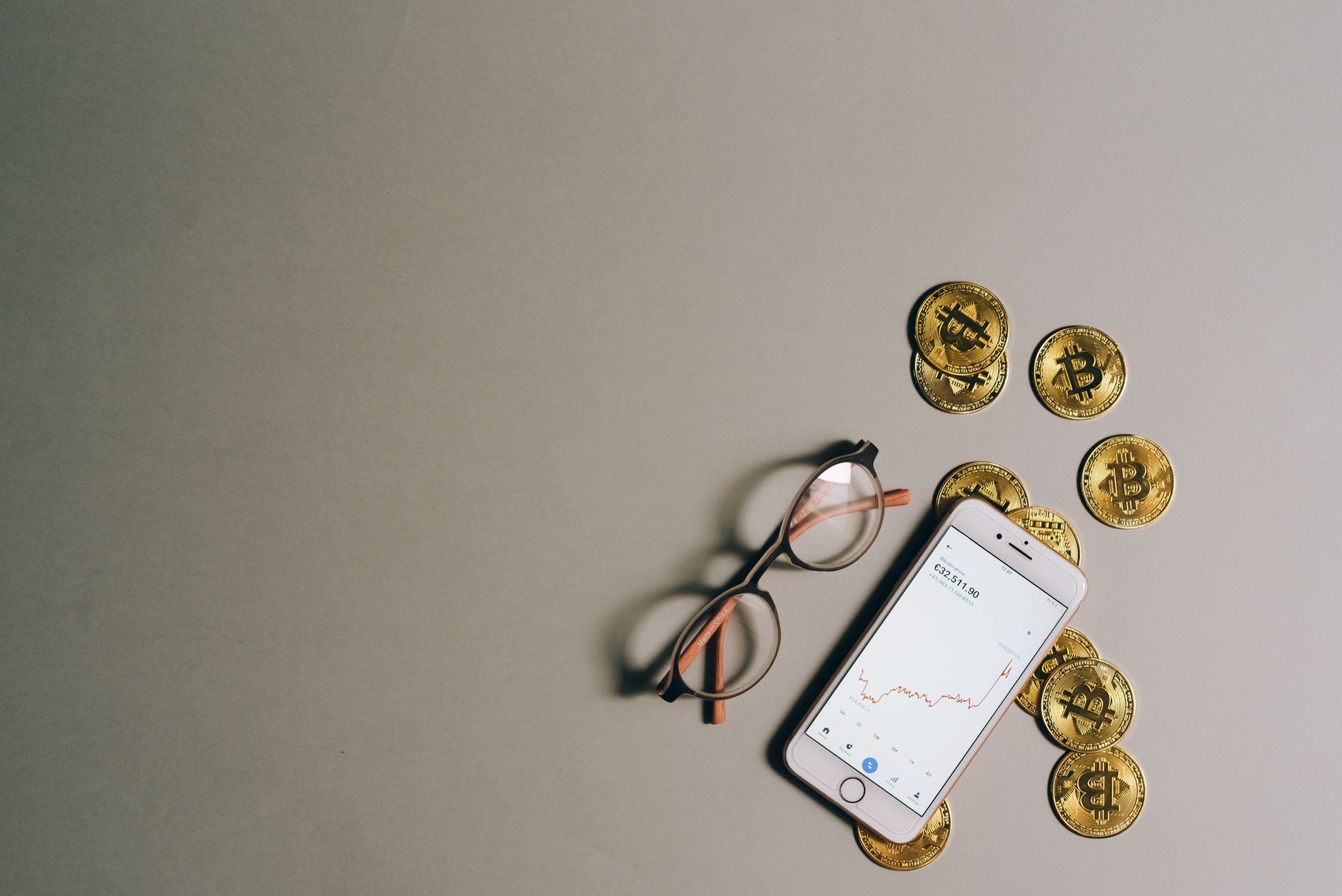 Flat lay of Bitcoin coins, smartphone with graph, and glasses on gray surface.