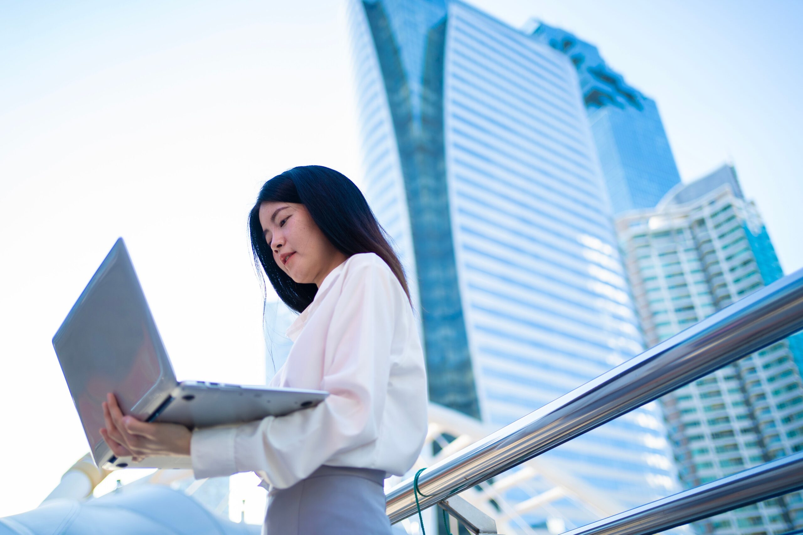 Confident Asian businesswoman working remotely on a laptop outdoors in Bangkok cityscape.