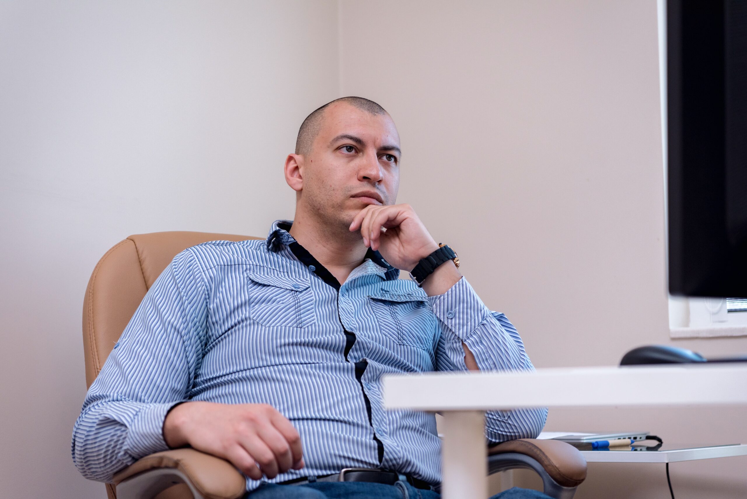 Man in striped shirt thinking while seated on an office chair indoors.
