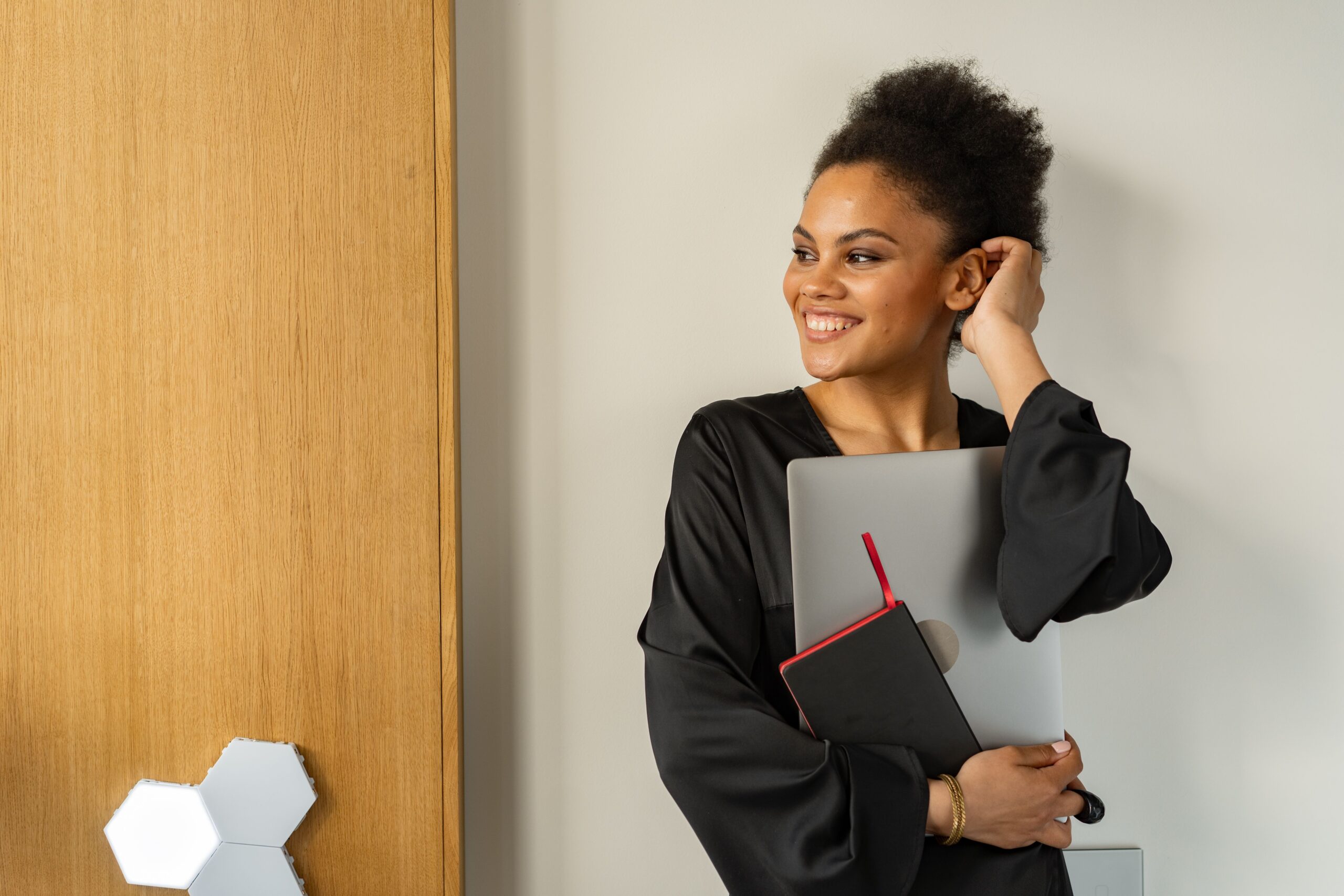 A confident woman with Afro hair smiling while holding a laptop and book indoors.
